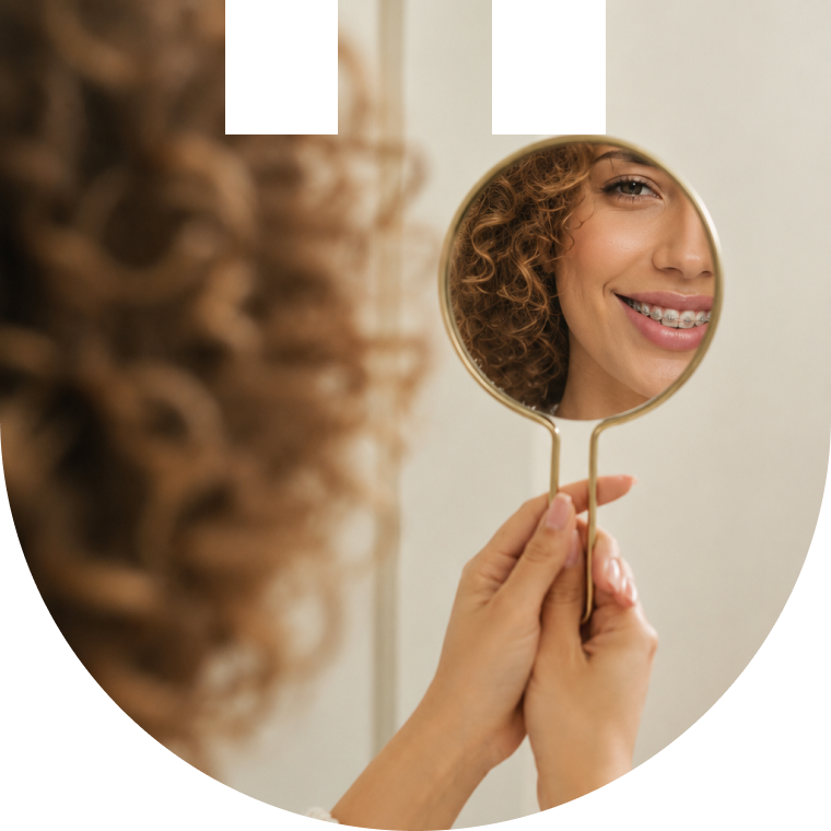 Patiente aux cheveux bouclés se regardant sourire dans un miroir à main, avec un appareil dentaire fixe